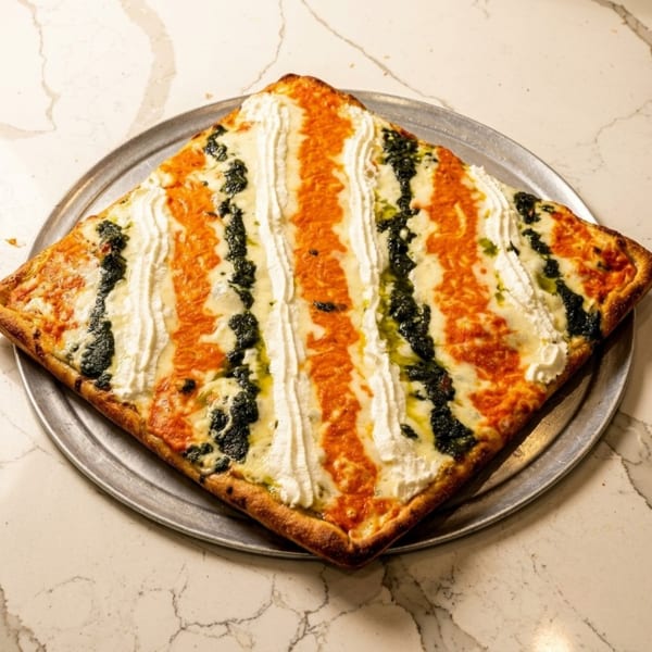 Square pizza with alternating stripes of red sauce, white ricotta, and dark pesto on a metal tray.